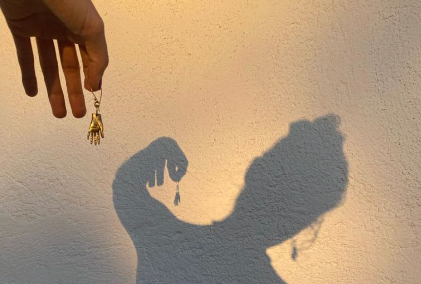 Golden Hour, Schatten einer Frau an einer Hauswand. Sie hält in einer Hand einen Ohrring der die Form einer Hand hat, den anderen trägt sie.