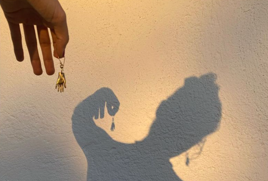 Golden Hour, Schatten einer Frau an einer Hauswand. Sie hält in einer Hand einen Ohrring der die Form einer Hand hat, den anderen trägt sie. 